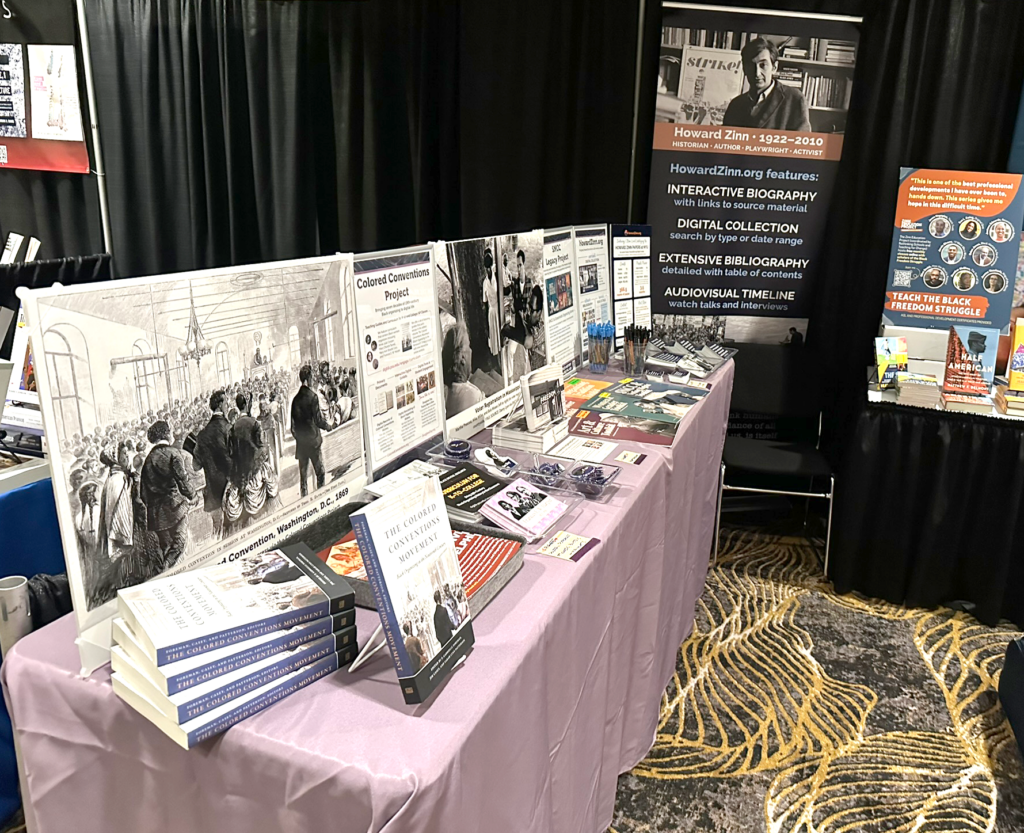Table with purple tablecloth and display of posters, books, and swag. Banner in the background.