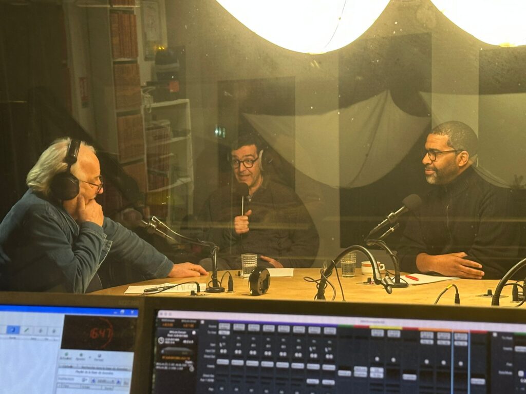 3 people with headphones on behind window in a radio studio